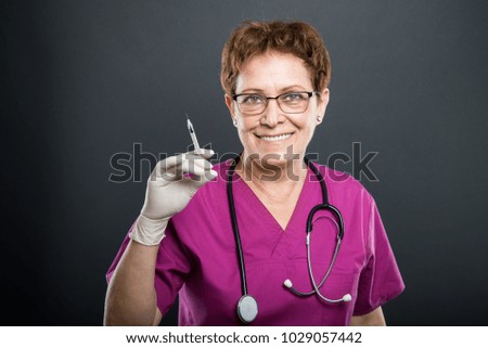 Portrait of senior lady doctor holding syringe and smiling on black background with copyspace advertising area