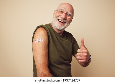 Portrait Of A Senior Male Smiling And Showing Thumbs Up After Getting A Vaccine. Mature Man With White Beard Sitting Against Brown Background Feeling Positive After Receiving Immunity Vaccination.