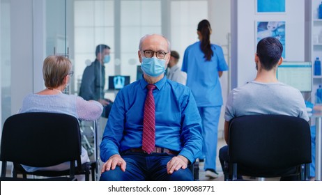 Portrait of senior man with face mask against coronavirus in hospital waiting area looking at camera. Patients during covid outbreak in clinic waiting for examination.
