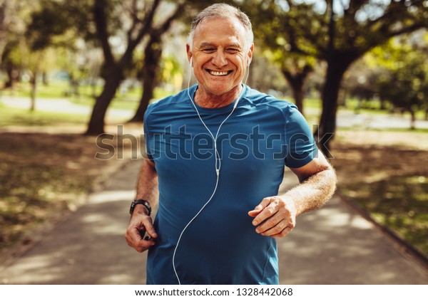 Portrait of a senior man in fitness wear\
running in a park. Close up of a smiling man running while\
listening to music using\
earphones.