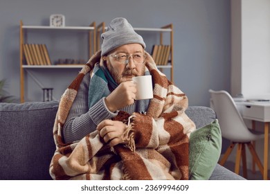 Portrait of a senior man in glasses sitting on a gray sofa at home wrapped in a blanket and in winter hat and trying to warm himself with a cup of hot tea. Heating problems concept.