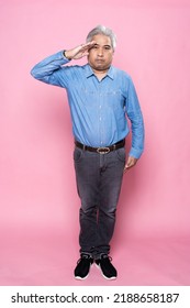 Portrait Of Senior Man Greeting The Camera With A Military Salute In An Act Of Honor And Patriotism, Showing Respect Against Pink Background, Happy Elderly Man Gray Hair In A Blue Shirt.