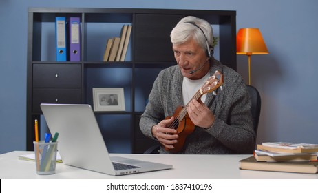 Portrait Of Senior Man In Headphones Taking Online Ukulele Lesson Looking At Laptop Screen. Retired Male Learning To Play Guitar Watching Webinar On Computer At Home