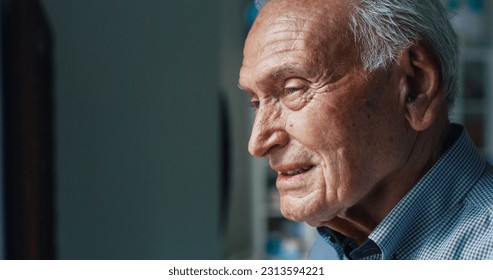 Portrait of a Senior Man at Home Getting Close to a Window and Watching Through it on a Sunny Day. A Smiling Old Man Watching his Grandchildren Play in the Yard. He is Enjoying the Warmth of the Sun