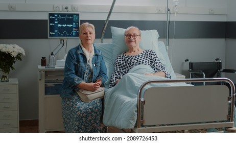 Portrait Of Senior Man In Hospital Ward Bed Receiving Assistance From Wife, Waiting On Medical Advice To Cure Sickness. Old Patient And Woman Getting Ready For Consultation At Clinic.