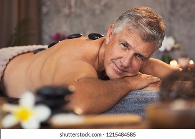 Portrait Of Senior Man With Hot Stones On Massage Table Looking At Camera. Smiling Mature Man Pampering Himself At Spa With Body Treatment. Man Lying And Relaxing During Hot Stone Massage.