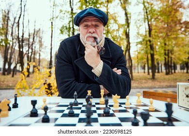 Portrait of senior man playing chess in the park on a daytime in fall. Thinking and smoking pipe. Concept of leisure activity, friendship, sport, autumn season, game, entertainment, old generation - Shutterstock ID 2230598125