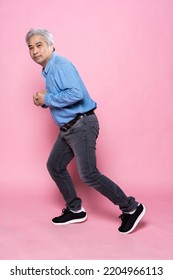 Portrait Of Senior Man Running Isolated On Pink Background, Happy Elderly Man Gray Hair In A Blue Shirt Black Jeans And Sneaker.