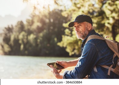 Portrait of senior man sitting near a lake with backpack and digital tablet. Mature male hiker sitting at a lake and looking away. - Powered by Shutterstock