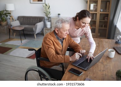 Portrait of senior man in wheelchair using laptop at retirement home with nurse assisting him, copy space