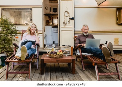 Portrait of senior married caucasian couple having rest on the deck chairs with food and wine just against their caravan home, celebrating special event while on a trip traveling by camper van - Powered by Shutterstock