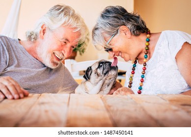 Portrait Of Senior Mature Caucasian People  Couple With Funny Pet Pug Dog Kissing And Having. Fun Together With Love And Happiness - Concept Of Animal Therapy For Aged Old Persons