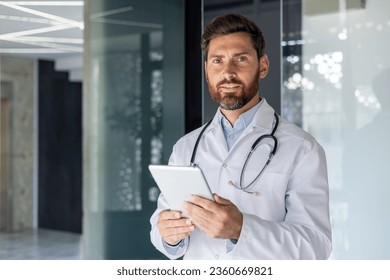 Portrait of senior mature experienced doctor, man in medical coat inside clinic looking at camera, senior doctor holding tablet computer, for online remote consultation of patients. - Powered by Shutterstock