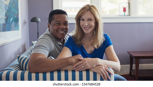 Portrait Of Senior Multi Ethnic Couple Sitting On Couch Smiling At Camera. Happy Black And White Husband And Wife At Home In Love With Beautiful House