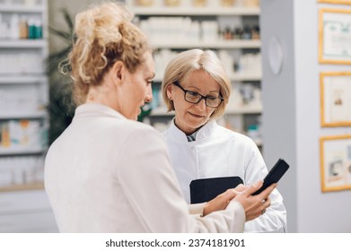 Portrait of a senior pharmacist woman healthcare worker working in a modern pharmacy and talking with her young colleague. Focus on a elderly female worker in a drugstore. Copy space. - Powered by Shutterstock