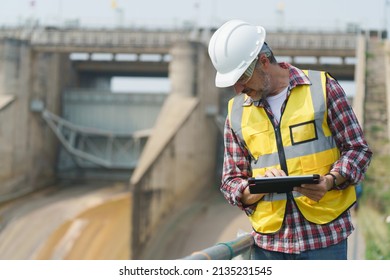 Portrait Of Senior Power Engineer Wearing Safety Jacket And Hardhat With Tablet Working At Outdoor Field Site Water Spillway Of Hydroelectric Power Plant Dam Electrical Generator At The Background.