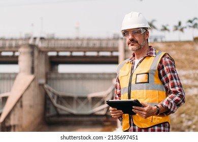 Portrait Of Senior Power Engineer Wearing Safety Jacket And Hardhat With Tablet Working At Outdoor Field Site Water Spillway Of Hydroelectric Power Plant Dam Electrical Generator At The Background.