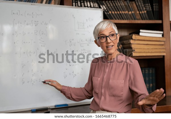 Portrait of senior professor explaining  math
formulas. Mature female teacher standing in classroom and talking
to her class. Lecturer explaining math formulas written on
whiteboard in library.