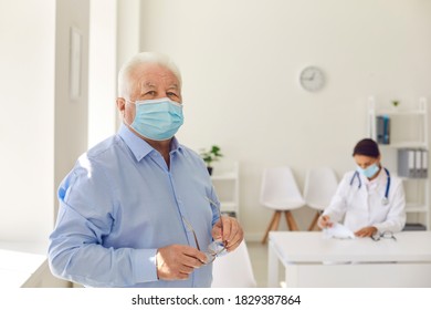 Portrait Of A Senior Retired Man In A Protective Medical Mask And With Glasses In His Hands On The Background Of A Doctor In A Hospital Office. Concept Of The Danger Of Coronavirus For The Elderly.