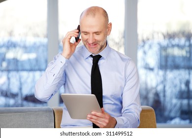 Portrait Of Senior Sales Man Sitting At Office While Holding Digital Tablet And Making Call. Businessman Analyzing Financial Data On Digital Tablet. 