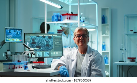 Portrait Of Senior Scientist Woman Looking Exhausted At Camera Sitting In Equipped Lab Late At Night. Team Examining Virus Evolution Using High Tech For Scientific Research, Vaccine Development