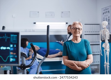 Portrait of senior woman with arms crossed waiting to exercise for physical therapy. Female patient with muscle injury looking at camera while preparing for leg physiotherapy on stationary bike. - Powered by Shutterstock