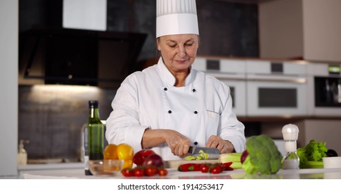 Portrait of senior woman chef cutting vegetables in modern kitchen. Professional female cook chopping onion on wooden boar preparing vegetarian dish with fresh vegetables - Powered by Shutterstock