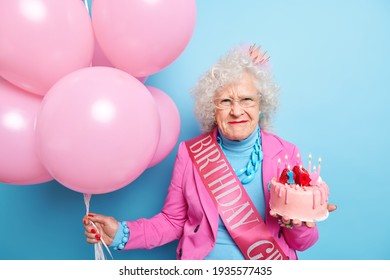 Portrait of senior woman with curly grey hair has dissatisfied face expression wears festive clothes celebrates birthday holds tasty cake bunch of inflated balloons isolated over blue background - Powered by Shutterstock