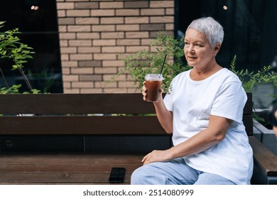 Portrait of senior woman drinking iced coffee at cafe, looking content and relaxed in city. European gray-haired older female sitting on bench, smiling happily, enjoying free time in retirement - Powered by Shutterstock