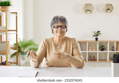 Portrait Of Senior Woman During Video Call, Educational Webinar, Web Conference Or Online Work Meeting. Happy Older Lady Sitting At Desk In Cozy Home Office, Holding Pen, Looking At Camera And Smiling