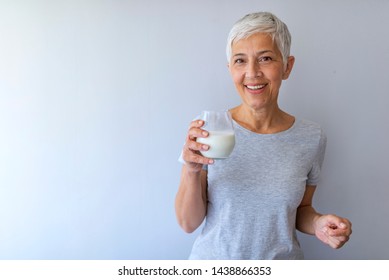 Portrait Of A Senior Woman With A Glass Of Milk. Beautiful Elderly Woman In Grey T-shirt With A Glass Of Milk On A Grey Background. Senior Woman Drinking Milk At Home