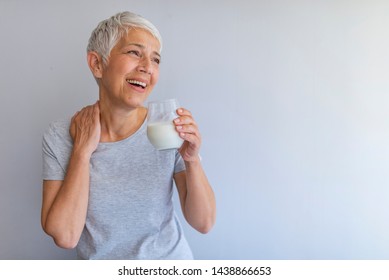 Portrait Of A Senior Woman With A Glass Of Milk. Beautiful Elderly Woman In Grey T-shirt With A Glass Of Milk On A Grey Background. Senior Woman Drinking Milk At Home