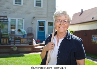 Portrait Of Senior Woman Holding Gardening Tool With Friend In The Background