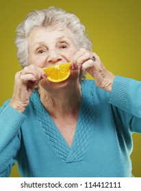Portrait Of Senior Woman Holding A Orange Slice In Front Of Her Mouth Over Yellow Background