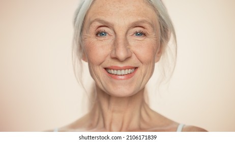 Portrait Of Senior Woman Looking At Camera, Smiling. Elderly Lady With Natural Grey Hair, Blue Eyes. Graceful Of Old Age Concept For Skincare, Beauty, Cosmetics Product With Isolated Beige Background