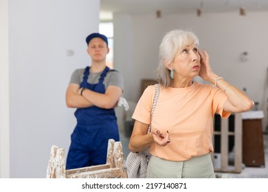 Portrait Of Senior Woman Making Helpless Gesture, Disappointed By Teenage Boy She Employed For Part-time Job In Construction Site. Young Man Wearing Overall, Standing In Background.