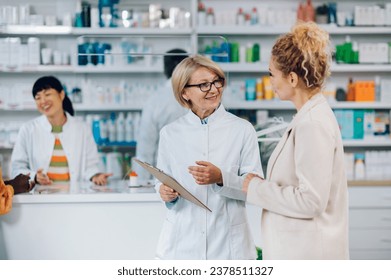 Portrait of a senior woman pharmacist working in a pharmacy while standing and talking with a patient. Elderly female worker wearing a white lab coat and talking to a customer in a drugstore. - Powered by Shutterstock