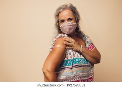 Portrait of a senior woman with protective face mask showing her arm with bandage after getting vaccine. Mature woman sitting against brown background after receiving corona virus vaccination. - Powered by Shutterstock