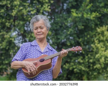 Portrait of a senior woman with short gray hair playing the ukulele while standing in a garden. Enjoy life after retiring. Concept of aged people and relaxation.