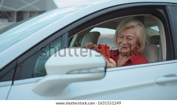 Portrait senior woman sits in car use red cell\
phone smile happy businesswoman blonde people smartphone cellphone\
communication internet search lady mobile bench browse network\
online slow motion