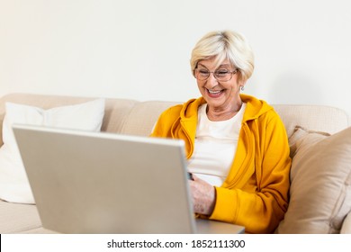 Portrait Of Senior Woman Sitting At Home On The Sofa And Working On Her Laptop. Older Lady Surfing The Net Or Having Video Call From Home And Feeling Exiciting. Holding Cup Of Coffee Or Tea