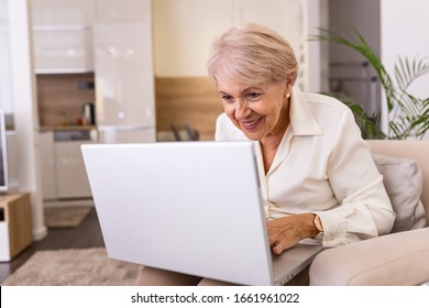 Portrait Of Senior Woman Sitting At Table At Home And Working On Her Laptop. Older Lady Surfing The Net From Home While Sitting On Her Sofa And Using Laptop Computer