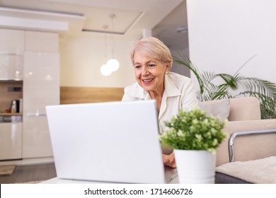 Portrait Of Senior Woman Sitting At Table At Home And Working On Her Laptop. Older Lady Surfing The Net From Home While Sitting On Her Sofa And Using Laptop Computer