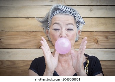 Portrait Of Senior Woman In T Shirt And Bandanna Showing Balloon Of Bubble Gum. Elderly Woman Having Fun Against Wooden Wall. Old Playful Woman Blowing Bubble Gum 