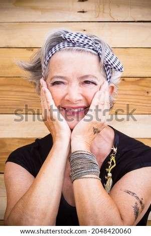 Portrait of senior woman in t shirt and bandanna with tattoo on hand. Cool elderly woman with hand on chin against wooden wall. Stylish old hipster woman with hands on cheeks