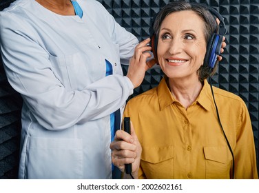 Portrait senior woman with white toothy smile while hearing check-up with ENT-doctor at soundproof audiometric booth using audiometry headphones and audiometer