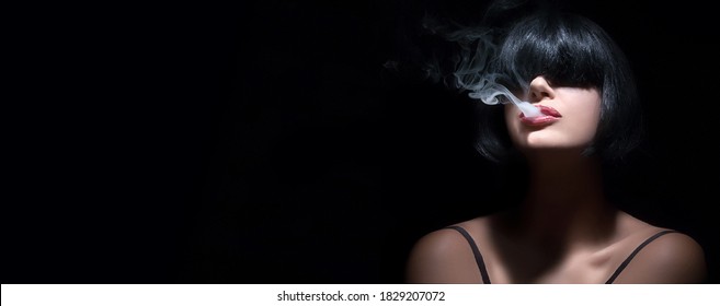 Portrait Of A Sensual Brunette Woman With Bangs Vaping Or Smoking As Smoke Curls From Between Her Parted Lips With Selective Focus And Shadows Over A Black Panorama Background With Copyspace