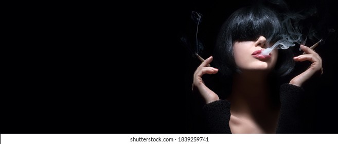 Portrait of a sensual brunette woman with bangs smoking two-handed as smoke curls from between her parted lips over a black panorama background with copyspace