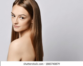 Portrait of sensual young slim woman with long silky straight hair standing with her back to camera looking over her naked shoulder at copy space over grey background. Haircare, beauty, wellness