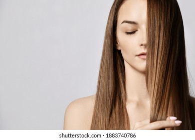 Portrait of sensual young woman with closed eyes and naked shoulders holding strand of her long silky hair covering half of her face over grey background with copy space. Haircare, beauty, wellness